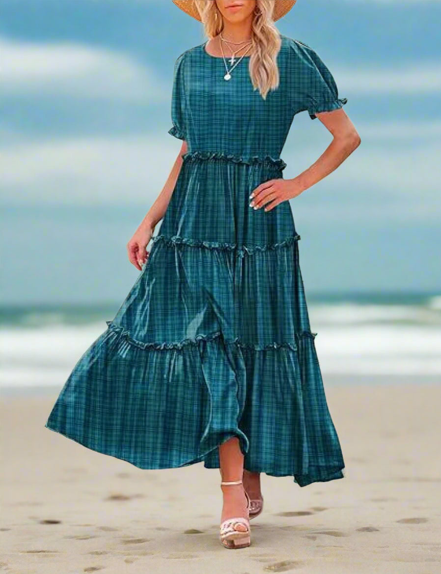 Woman wearing a teal plaid dress standing outdoors with a stone building and plants in the background.