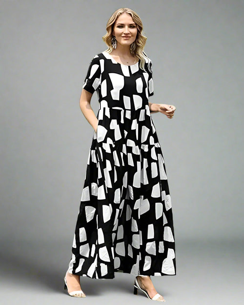 Woman wearing a black and white patterned dress standing outdoors on grass with trees and a white fence in the background.